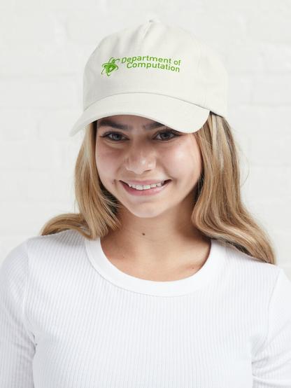 Photo of a woman smiling while wearing a white hat with the green Department of Computation word logo, which is a green silhouette of a Quokkium atom, which in turn is a quokka's head surrounded by three atomic orbits, with a lightning bolt symbol in the middle. The name of the titular department is to its right.