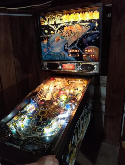 A Data East Time Machine pinball. It's glowing in a darkened room. The backglass shows a light blue car with people representing stereotypical people from the 80's, 70's, 60's, and 50's in (or hanging off of) it. The machine has four alphanumeric displays to show the scores and information.