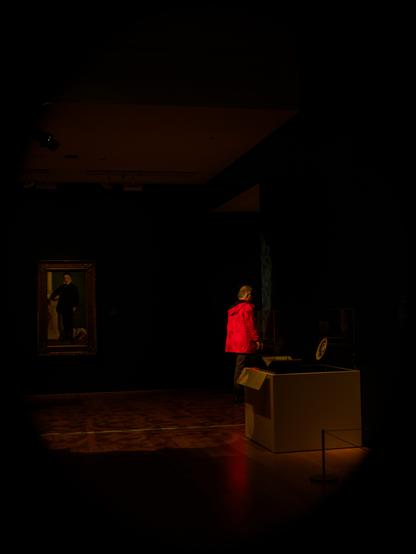 Low key mostly black photograph of a painting at the Art Gallery of Ontario. The painting is in the background on the left. Within the shadows, a man wearing a red coat is walking to the right.