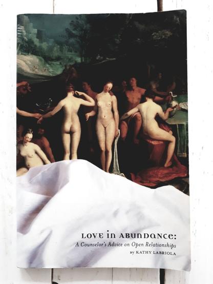 The book "Love in abundance - a counselor's advice on open relationships" laying on a white table. It is a paperback book with a historical painting by Karel van Mander. The painting shows a couple of naked people in a typical 17th century setting. The female nudes are in light colours at the front and the male nudes are in darker colours at the back. They seem to be having a party (instruments and carafes) in the countryside. The painting is half covered by a white curtain. The title is in the right-hand corner of the cover. Also the name of the author, Kathy Labriola.