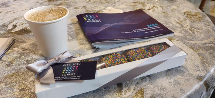A rectangular white box with a ribbon bow, containing multicoloured sprinkle-covered Oreos. It's on a swirly gold and grey tablecloth, with a cup of coffee and a conference booklet next to it.
