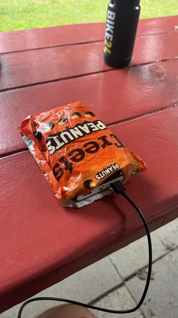 Ein zerknittertes orangefarbenes Treets-Pack mit einem USB-Kabel, das darauf angeschlossen ist und auf einem roten Picknicktisch ruht. Eine schwarze Wasserflasche mit dem Text "BIKE24" befindet sich im Hintergrund.