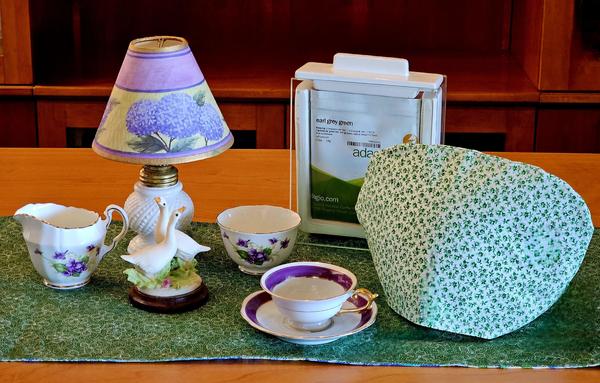 A tea tableau in green & purple, with touches of white like a small hobnail milk glass lamp & a graceful bisque figurine of geese. The tea is Earl Grey Green tea from Adagio.