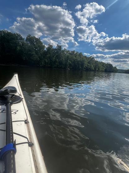 View of the Schuylkill River, with the bow of a white kayak along the left side of the view. The river, lightly rippled and brightly reflecting the sky, runs off to the right, and a treeline beneath a lightly cloudy sky crosses the view just above the halfway point.