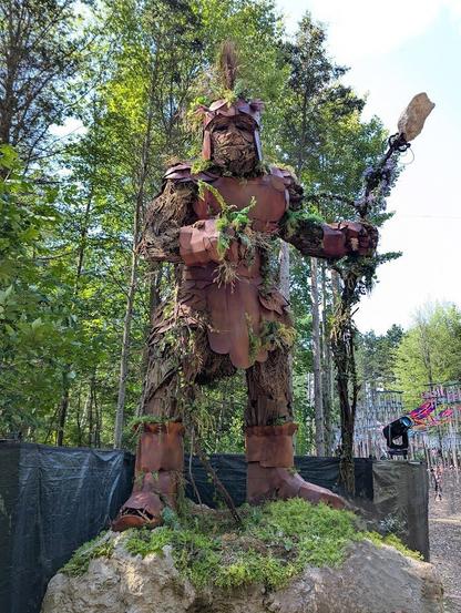 A sculpture of a large person in armor made of wood and bark. He has a large walking stick with a crystal at the end and is standing on a large rock.