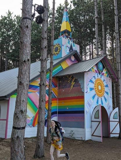 A large chapel is built in the middle of the woods from reclaimed material.  It's doors are open to all and it is adorned with bright painting featuring a sun, storm, rainbow and trans flag colors. A festivalgoer with long brown hair, a multicolored top and yellow skirt is running by.