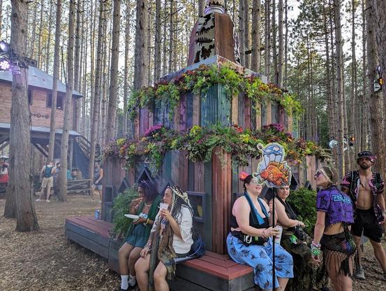 Multiple festivalgoers sit in front of a multi tiered planter made of various colors of wood with bright and vibrant plants growing from every tier. In the background a raised cabin type building is surrounded by more people.