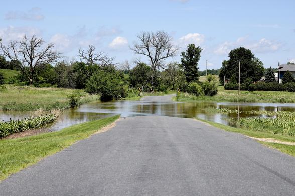 Morill Road closed by high water after recent rain. Road view with water across before a bridg and the. Oak tree near a road bend to the right. Blue sky and clouds