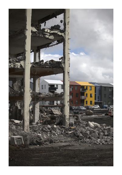 A partially demolished building stands in the foreground with exposed concrete, steel rebar, and piles of rubble at its base. In the background, modern apartment buildings painted in bright colors such as yellow, blue, and red are visible under a cloudy sky. The area around the demolished structure appears desolate, with scattered debris and construction materials.