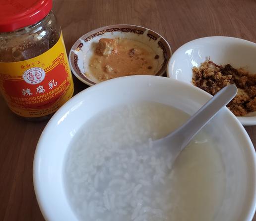 Bowl of plain rice congee with Chinese soup spoon in it. In front of my congee bowl, left to right:
Glass jar of Fat Choy Brand preserved chili bean curd, small sauce dish with some of the bean curd in it & dried shredded pork with soya sauce in small sauce dish.