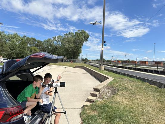 Two children are sitting in the open trunk of a car, using a telescope to look at an airplane flying overhead.