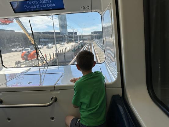 A child in a green shirt looks out the front window of a moving train, observing the tracks and the cityscape ahead.