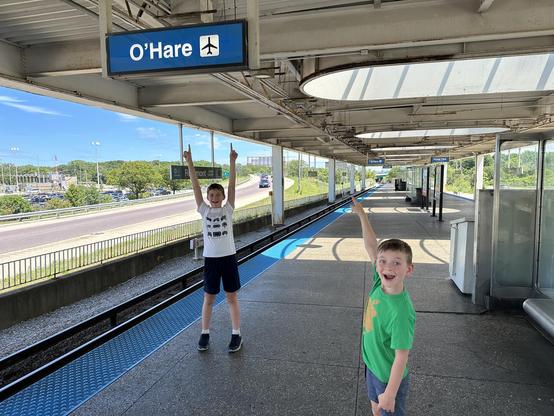 Two children smiling and pointing upwards at an "O'Hare" sign at a train station platform. The station has a covered roof with exposed beams and a view of a road and greenery in the background.