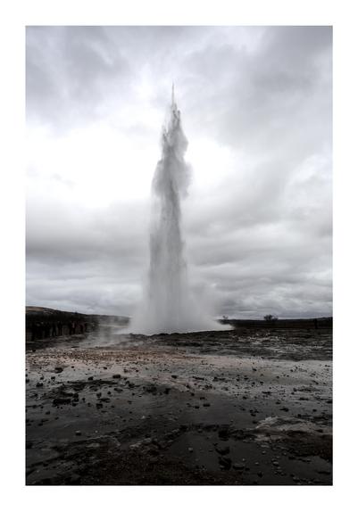 A powerful geyser, known as Strokkur, erupts in a dramatic plume of water and steam against a cloudy sky. The eruption reaches high into the air, contrasting with the dark, rocky landscape below. In the distance, small figures of onlookers can be seen observing the natural spectacle, emphasizing the geyser's immense size and force. The surrounding area is wet and rugged, typical of geothermal regions.