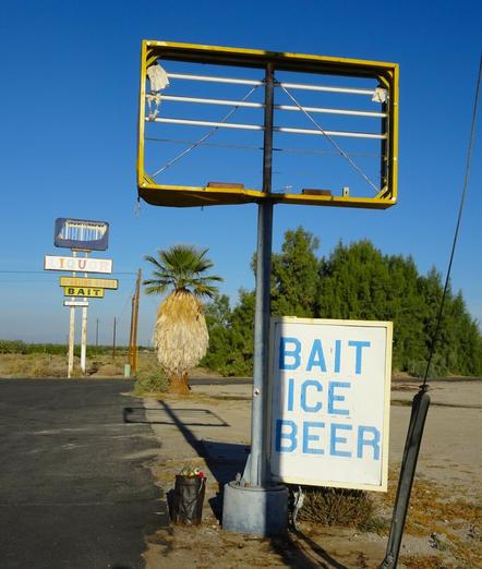 Against a blue sky on a sunny day, old signs on an abandoned property that used to be a minimart and/or gas station. In the foreground, a tall pole supports a rectangle metal frame that used to have a lighted sign in it. Beneath that, and attached to the pole is a white sign with faded blue letters that say "Bait, Ice, Beer." In the background is another vintage midcentury modern collection of signs: an empty frame, one that says "liquor" in a cheerful, funky midcentury font, and two other signs that say "Sporting Goods" and "Bait." There is a palm tree next to the asphalt parking lot. In the distance, a tuft of green trees.