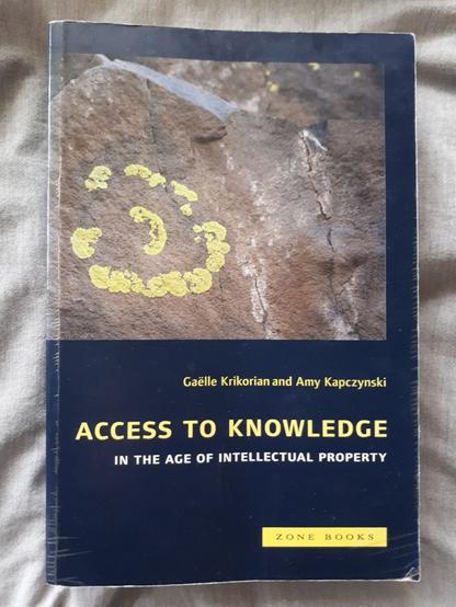 The book "Access to Knowledge" on a grey curtain. It is a thick black paperback. On the cover is a close-up of a grey stone with some kind of yellow moss in a circle. The moss looks a bit like the copyright symbol. At the bottom of the page, in yellow letters, are the title of the book, the editors (Gaëlla Krikorian and Amy Kapczynski) and the publisher (zone  books).