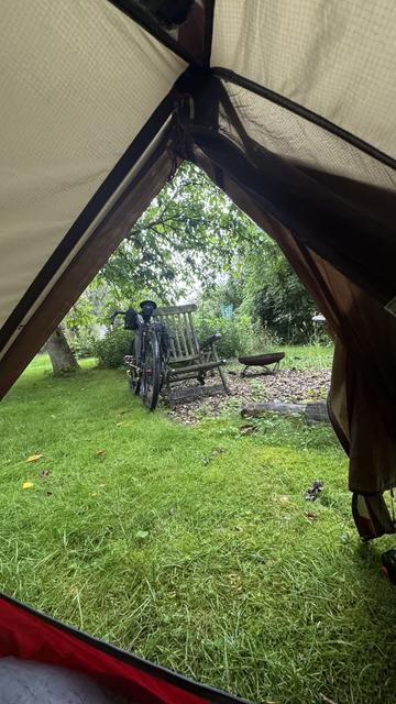 Blick vom Inneren eines Zelts mit Blick auf eine Grasfläche, mit einem Fahrrad, das teilweise neben einer Holzbank und Bäumen im Hintergrund sichtbar ist.