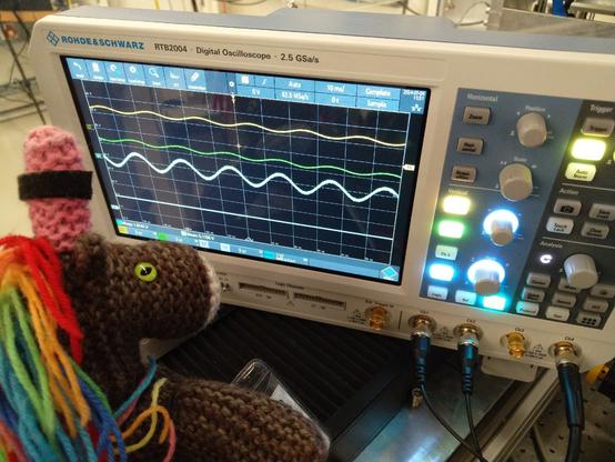 Max Horn, das bunte Strickhorn, betrachtet konzentriert das Display eines Digitaloszilloskops der Marke Rohde & Schwarz RTB2004. Auf dem Bildschirm sind zwei differentielle Signalkurven (gelb und grün) sowie deren Differenzkurve dargestellt. Die Signale stammen vom Motorencoder des Röntgenmikroskops. Die Amplitude der Kurven beträgt etwa 1 Volt, was den Erwartungen entspricht. 

Zusätzlich zeigt das Display eine Versorgungsspannung von ca. 5,1 Volt an, was darauf hinweist, dass die Elektronik ordnungsgemäß funktioniert. Max scheint stolz auf die erfolgreiche Reparatur des Röntgenmikroskops zu sein, die er durchgeführt hat. Die detaillierte Analyse und Darstellung der Signale auf dem Oszilloskop verdeutlichen den technischen Erfolg und die Präzision der durchgeführten Arbeiten.