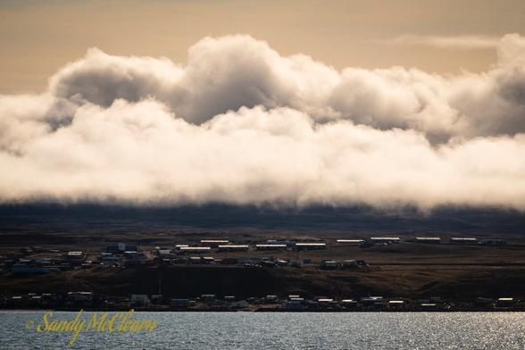 A thick layer of cloud appears to hang over sunlit rows of buildings on the shoreline.
