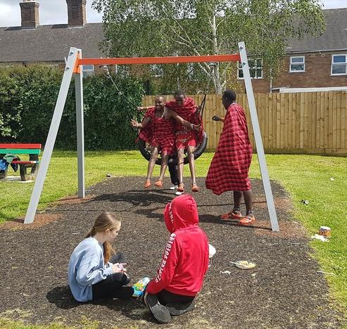 Two Maasai men in traditional dress try out the park's swing, as a third man and two young participants look on.