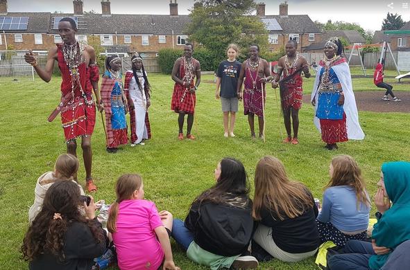 In a park in Chipping Norton, the Kenyan visitors, dressed in traditional red checked clothing and beaded jewellery, share information about their culture and daily lives as they stand in front of a group of seated young people involved in Transition Chipping Norton's youth project.