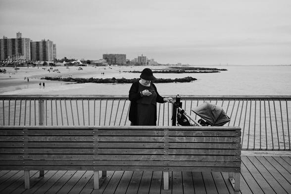 A person stands near a pram on Coney Island boardwalk overlooking the beach, with the pier and summer visitors in the background.