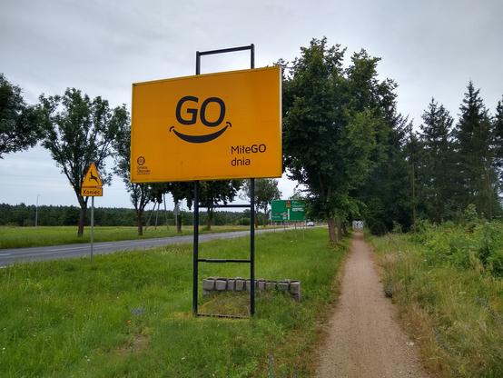 Billboard wzdłuż drogi. Na żółtopomarańczowym tle umieszczono uśmiechniętą "buźkę", składającą się z nieco pochylonych liter GO i łuku przypominającego usta. W lewym dolnym rogu miśeci się logotyp segmentowanego okręgu z literami "GO" w środku, a pod nim napis "Gmina Oborniki", "paleta możliwości". W prawym dolnym rogu duży napis "Miłego dnia", w którym "GO" napisano kapitalikami.