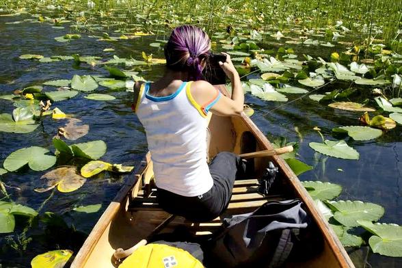 Photo from my friend.
Perspective from back of canoe of my back, photographing water lilies & lilypads, from front of canoe. I'm wearing tank top & black pasta. A canoe paddle is laying across my legs. My yew wood cane is below my seat.