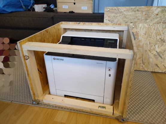 Ein weißer Laserdrucker steht in einer Box aus OSB-Platten.