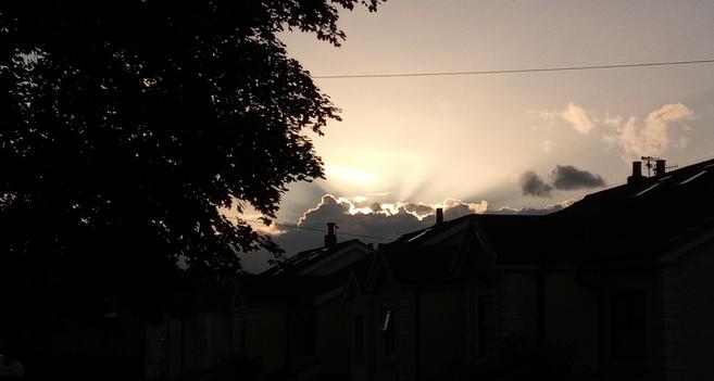 The rays of the sinking sun breaking through a cloud over a line of roofs in Disley, with tree to left.