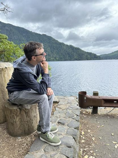 A white man wearing sunglasses sits in The Thinker pose on a stump, looking out over a blue forest mountain lake with a cloudy grey sky above.