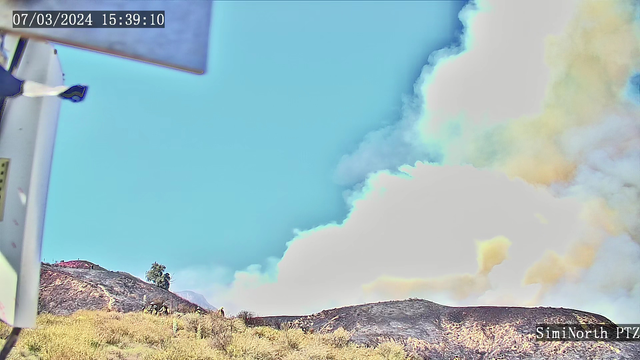 Photo from a site with brush in the front and a fire and a black hillside not far away