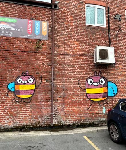 Streetartwall. A mural with two funny fat bees has been sprayed/painted on a red brick wall. The simply designed pair have an oval body, blue wings, two big eyes and a smiling mouth with teeth and no nose. The two cheerful bees are now waving to drivers in a parking lot and spreading good cheer.