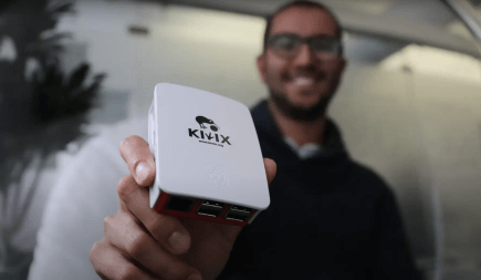 The man smilingly holds a very small computer with the Kiwi logo on it in his hand.