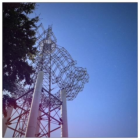 Metallic abstract sculpture against a clear blue sky, partially framed by tree branches on the left.