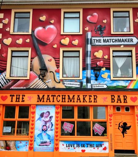 Ein bunt angemaltes zweistöckiges Haus, unten orange und oben rot, mit vielen kleinen und großen bunten Herzen, Fensterrahmen und einem Regenbogen. Unten steht groß "The Matchmaker Bar" und oben gibt es einen Wegweiser, auf dem ebenfalls "The Matchmaker" steht.

A brightly painted two-storey house, orange at the bottom and red at the top, with lots of small and large colourful hearts, window frames and a rainbow. "The Matchmaker Bar" is written in large letters at the bottom and there is a signpost at the top that also says "The Matchmaker".