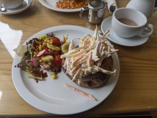 A white plate with a big baked potato, cut in length and filled with coleslaw. The second half of the plate is occupied by a small side salad.
Next to the plate there is a cup of tea on a saucer and a white pot. Also: There's a small stainless steel cup with milk.
