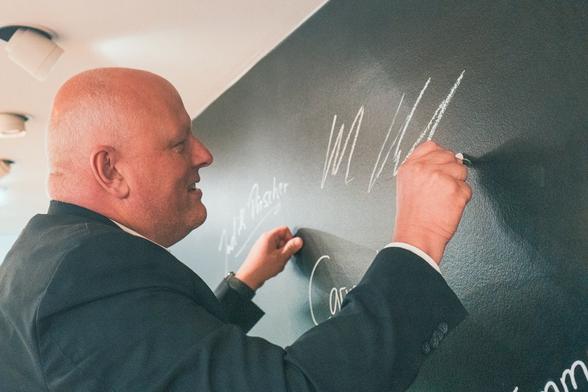 Ulrich Kelber schreibt an den oberen Rand einer Wand, die mit einer Tafeloberfläche versehen ist, mit Kreide seine Unterschrift
