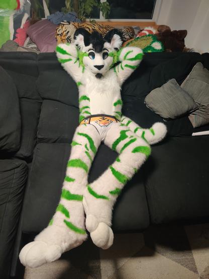 A white tiger fursuit with green stripes lies on a couch, the camera looking down at him from above. Both his hands are held behind his head. His eyelids are partially shut, giving him "bedroom eyes". He's wearing a pair of underwear with a tiger face on it.