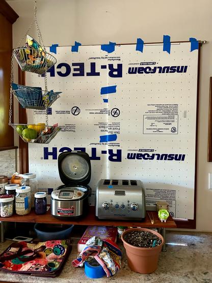 A kitchen wall. Two foam “R-TECH INSULFOAM” panels black a window and are held in place with blue tape. You can see a set of 3 hanging, wire baskets with citrus fruits in the bottom basket. A stone countertop has a flower pot, shopping bag, and the blue tape roll. There’s a shelf on the counter with a toaster, rice cooker with an open lid, jars of breakfast cereal, and issue ids and ends.