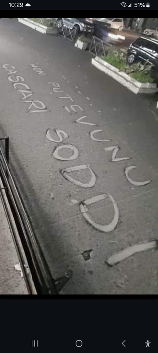 Un marciapiede coperto di cenere vulcanica in cui qualcuno ha “pulito” due righe di testo che recitano: «nun putevunu cascato soddi» (non potevano cadere soldi)