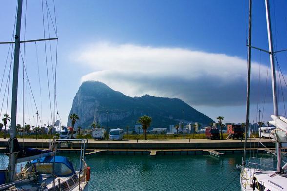 View of Gibraltar from the marina of La Linea, which is just on the other side of the border in Spain. The rock of Gibraltar, jutting out of the flat surroundings is crowned by a cloud that shows the shape of the updraft on the eastern side where it is created and extends darkly over the west where most of the settled part lies. The surrounding weather is clear blue skies. A few boats are docked in the marina, framing the shot.