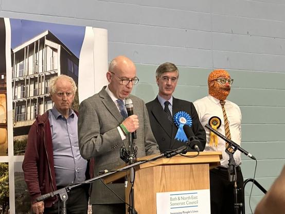 Jacob Rees Mogg, a man whose face one would never tire of punching, is standing on stage at the vote count, next to a more serious candidate.