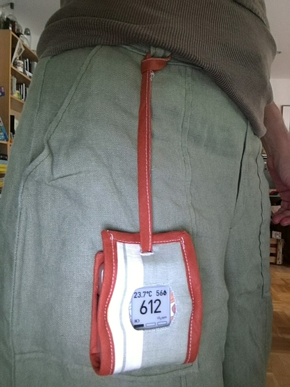 An Aranet4 inside a cloth cover is hanging from a belt loop. The belt loop is on some green linen pants. The pants are very creased because they are linen.

The cover has vertical green and cream colored stripes and its binding is a terracotta brown.