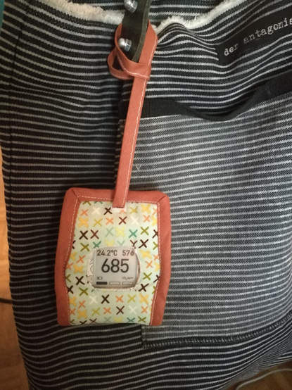 This is the same Aranet4 but in another cloth cover. This time the cover is hanging from the strap of a handbag. The handbag has black and white horizontal stripes.
The cloth cover for the Aranet4 has the same terracotta brown binding as the other one but this time the outer fabric is light blue with brown, green, yellow, and white Xs on it.