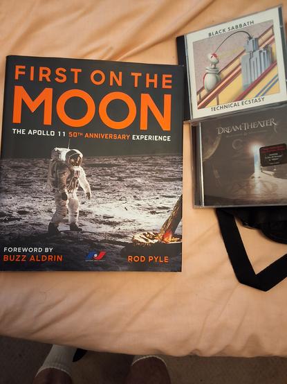A hardcover book called "First On The Moon" by Rod Pyle, and two CDs: Black Sabbath's Technical Ecstasy and Dream Theater's Black Clouds & Silver Linings.