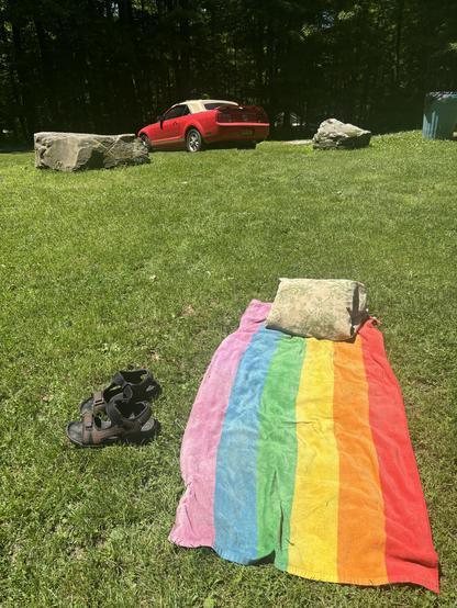 A rainbow towel laid out on the grass, a pillow, on the side a pair of sandals. In the distance a red car with tan convertible roof is parked.