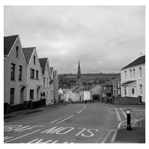 a black and white photo of a two lane road running between some houses, downhill. In the distance is a church spire, and behind it some countryside hills