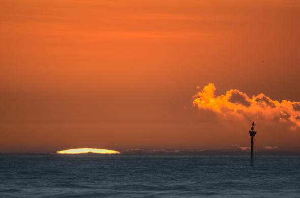 A seafront view of the tip of the sun rising in the horizon with an almost golden sky above. A navigation beacon can be seen in the foreground bottom right corner composing with a tiny cloud over it.