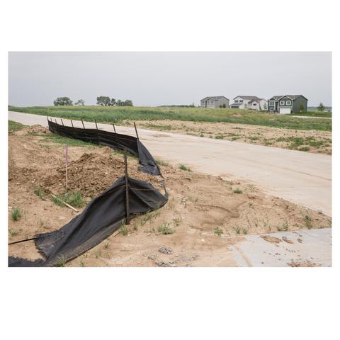 Three new homes sit in the upper right corner of the image in a new lot. A new road bisects the image diagonally through the center. Grass separates the homes from the road. A temporary fabric fence lines the road, facing the three houses in the distance.